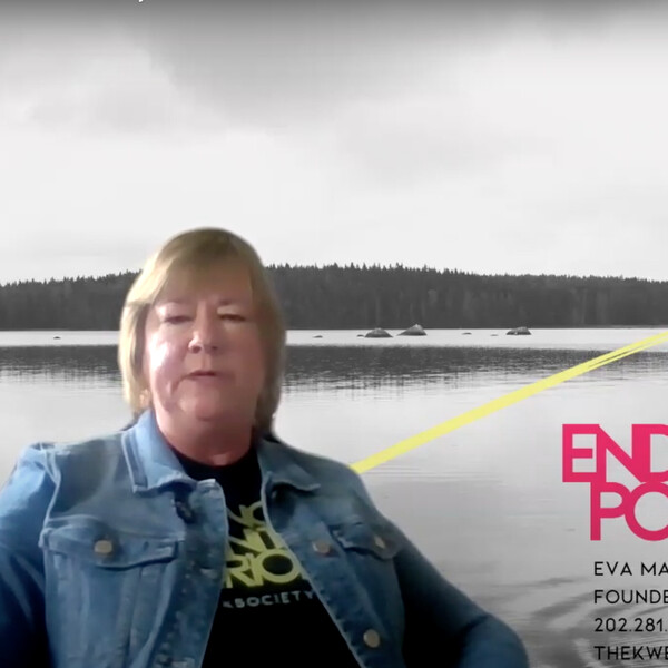 An Indigenous woman stares at the camera, she's wearing a denim blazer, she's in front of a virtual background that says the Kewk society and shows a lake with trees.