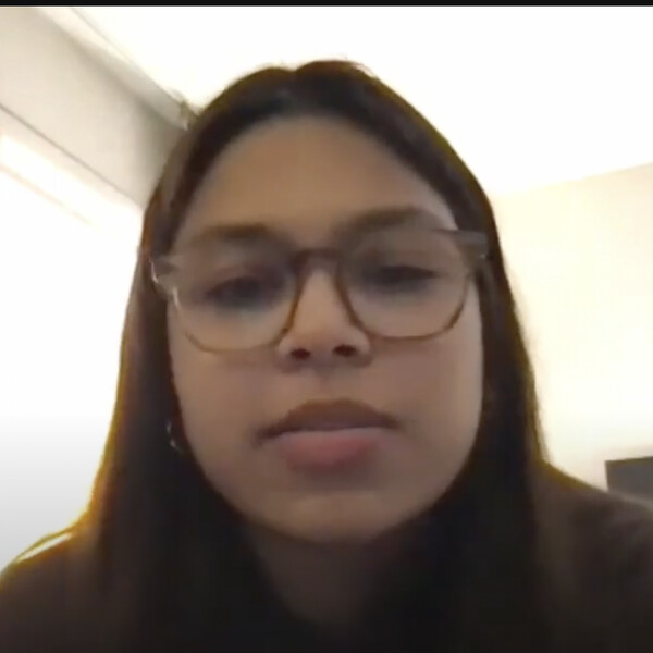 Isabella looks at the camera, and she is wearing eyeglasses and a dark shirt. She sits in a room with a bright window and tan walls.