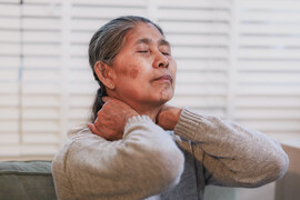Senior woman at home holding her neck with eyes closed, showing pain, stiffness, stress or fatigue. Warm indoor portrait with soft light and calm background.