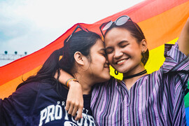 Two women hugging in front of rainbow flag.