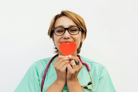 Woman in green scrubs with stethoscope around her neck holds red paper heart in front of her face.