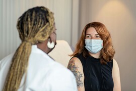 A doctor with a face mask talking to a seated patient.