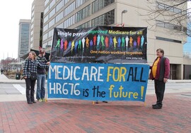 A group of people stand next to banners that read: "Single Payer: Improved Medicare for All. One nation working together. Physicians for a National Health Program." and "Medicare for All, HR676 is the future!"