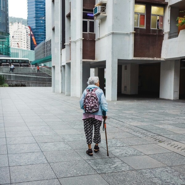An elderly woman in bright colors and patterned clothes walks across a square in Hong Kong, beside an apartment block. She holds a walking stick. A busy urban environment and traffic is visible in the distance.