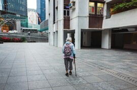 An elderly woman in bright colors and patterned clothes walks across a square in Hong Kong, beside an apartment block. She holds a walking stick. A busy urban environment and traffic is visible in the distance.