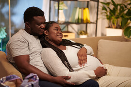 A Black man and a pregnant Black woman sit together on a couch.