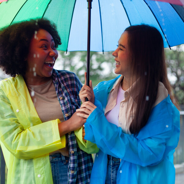 Two women are standing under a rainbow umbrella, smiling and laughing.