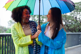 Two women are standing under a rainbow umbrella, smiling and laughing.