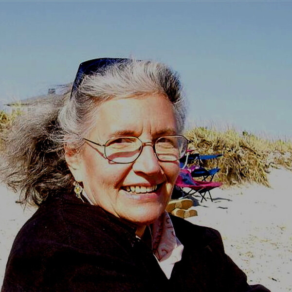 color photograph of Norma Meras Swenson, smiling, wearing glasses and a black top, ger gray hair swept back by the wind, in front of sand dunes, under a blue sky.