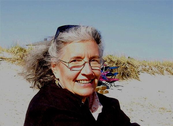 color photograph of Norma Meras Swenson, smiling, wearing glasses and a black top, ger gray hair swept back by the wind, in front of sand dunes, under a blue sky.