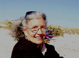 color photograph of Norma Meras Swenson, smiling, wearing glasses and a black top, ger gray hair swept back by the wind, in front of sand dunes, under a blue sky.
